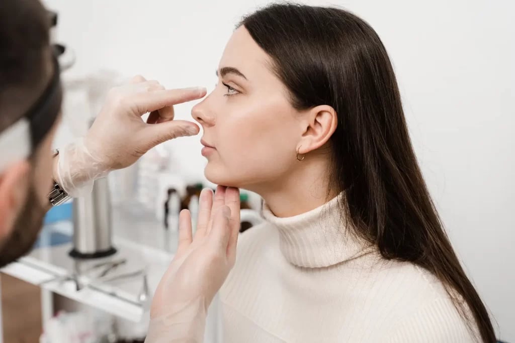 Medical professional examining a woman's nose and chin profile while she sits upright during a consultation.